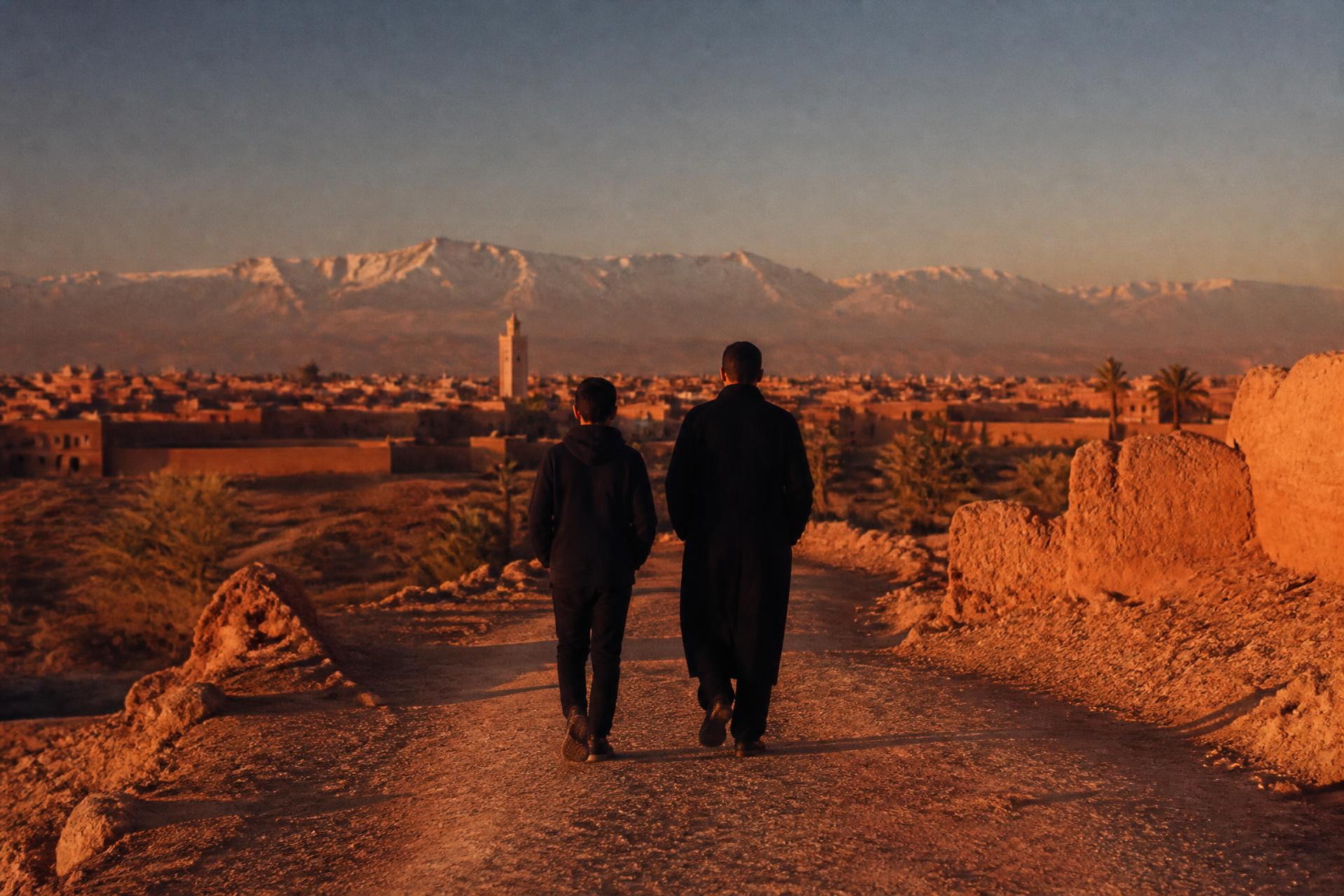 Un éducateur et un adolescent marchent de dos sur un chemin de terre vers les montagnes de l’Atlas, aux abords de Marrakech